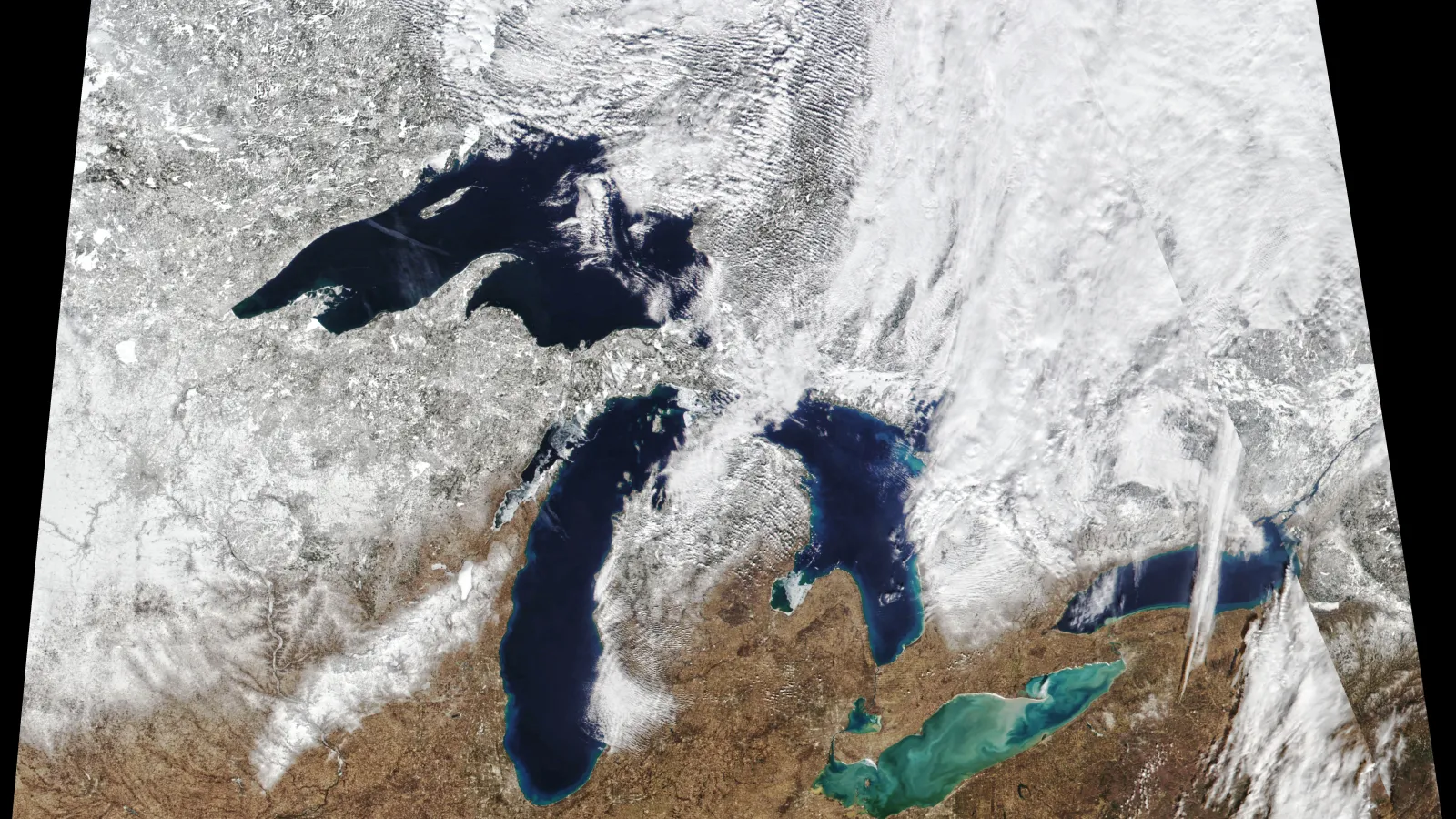 Satellite image of the great lakes with the top half of the image white, covered in snow and the bottom brown and the lakes dark and light blue.. 